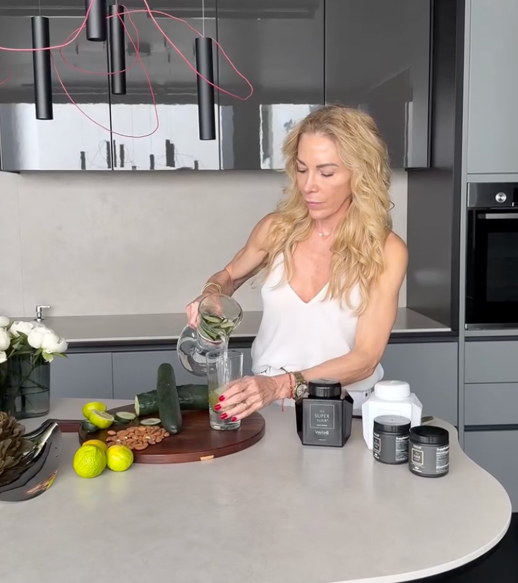 A woman in a modern kitchen pours water infused with cucumber slices from a pitcher into a glass.